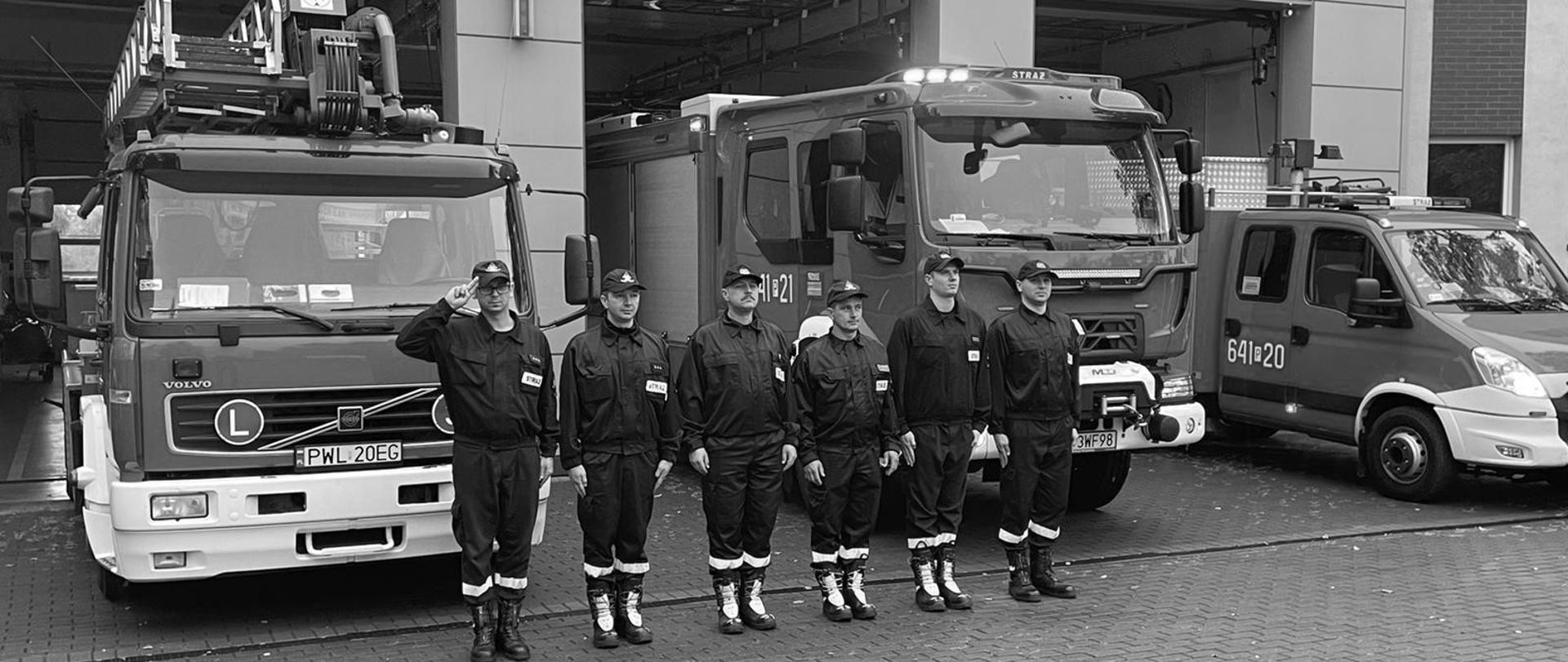 Na zdjęciu widoczna zmiana służbowa JRG Wolsztyn oddająca hołd zmarłemu druhowi. Na drugim planie samochody pożarnicze z włączonymi sygnałami świetlnymi i dźwiękowymi. W Tle komenda powiatowa PSP w Wolsztynie. Zdjęcie w szacie czarno-białej na znak żałoby