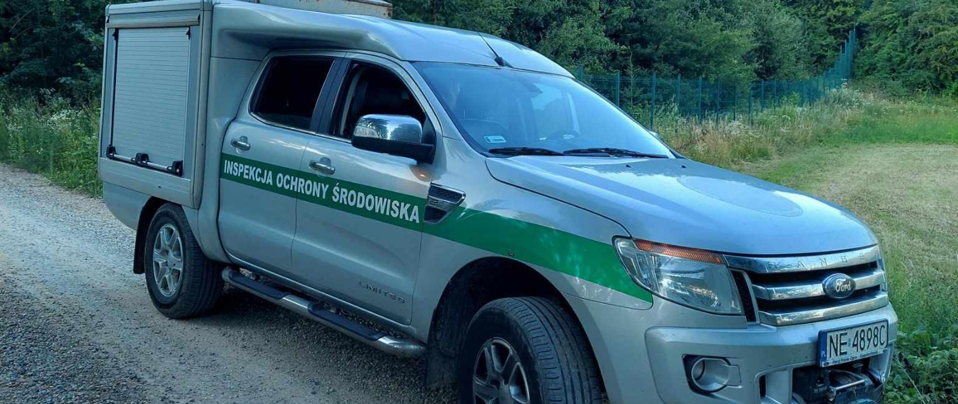 Samochód terenowy szary z zielonym paskiem pośrodku, na którym jest biały napis Inspekcja Ochrony Środowiska. W tle łąka, płot i las.