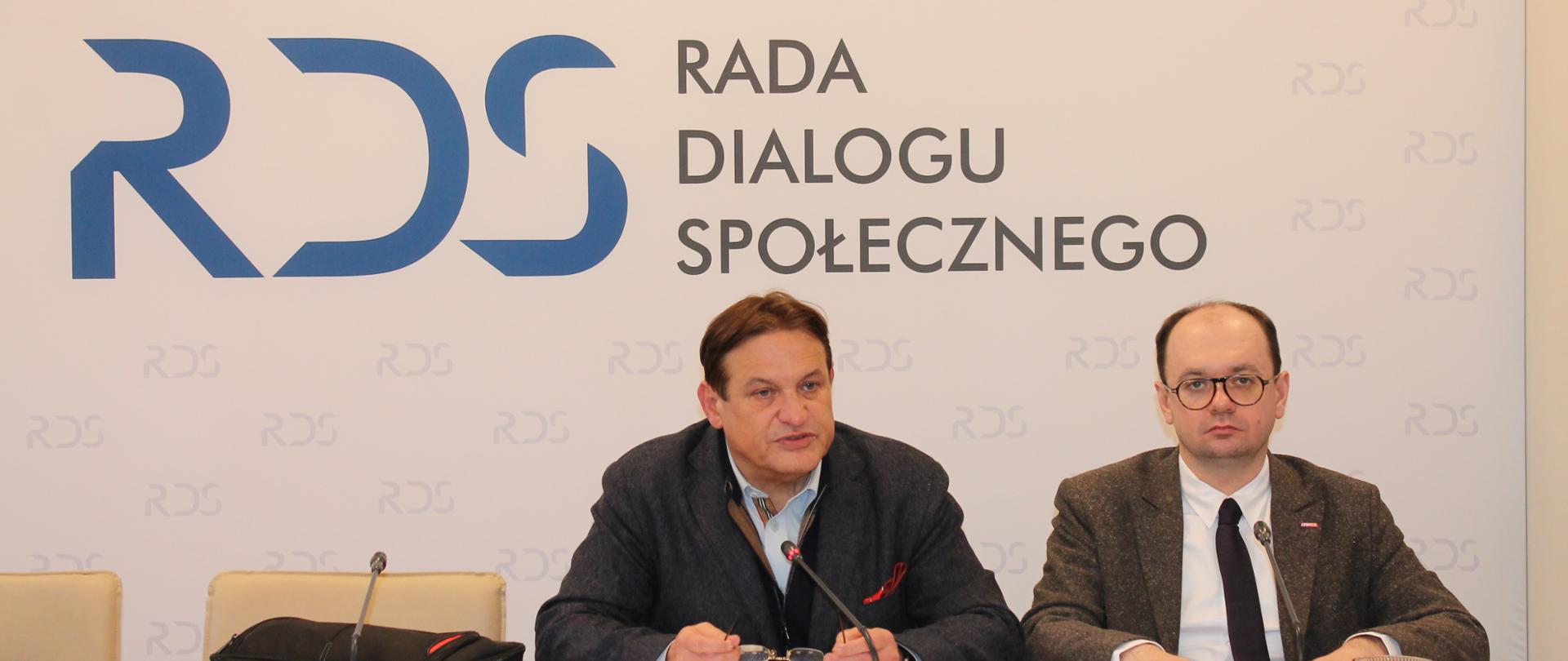 Zdjęcie przedstawia stół prezydialny za którym siedzą: Pan Jacek Męcina, Przewodniczący Zespołu oraz Pan Sebastian Gajewski, Podsekretarz Stanu w Ministerstwie Rodziny, Pracy i Polityki Społecznej. W tle znajduje się baner z napisem Rada Dialogu Społecznego.