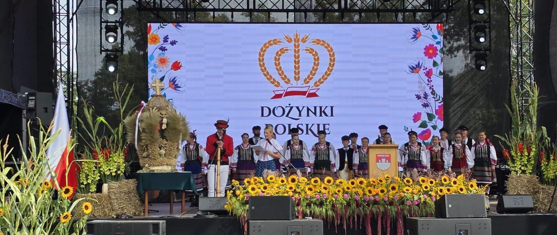 Na scenie plenerowej udekorowanej wieńcami , słonecznikami i kwiatami występuje zespół w strojach ludowych.