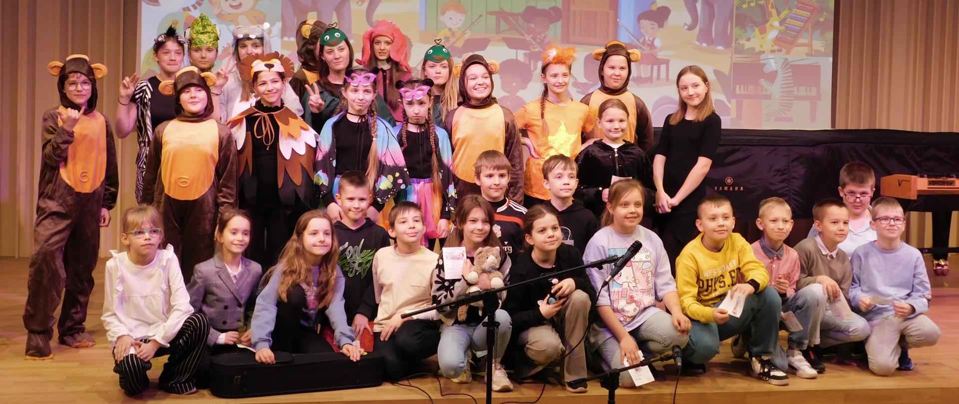 Na zdjęciu duża grupa uczniów. Część z nich przebrana za zwierzęta. w Tle za nimi na rzutniki domek, a wokół niego zwierzęta. W górnej części czarny napis: "dziękujemy za przybycie. zapraszamy do naszej szkoły".