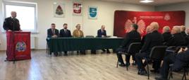 Strażak w mundurze wyjściowych przemawia do zebranych na sali osób, ze stołem prezydialnym siedzi kobieta i trzech mężczyznm, przodem do nich siedzą starsi wiekiem mężczyżni