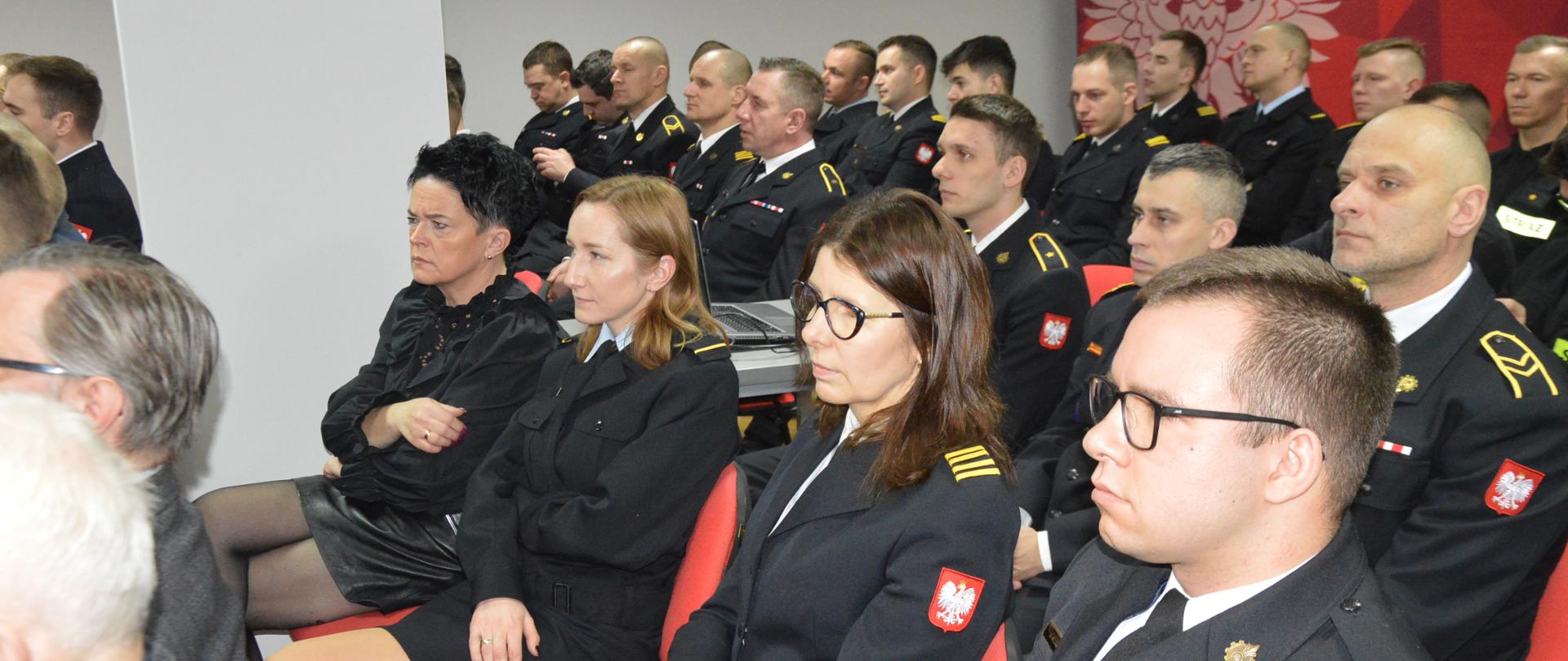 Narada roczna. Świetlica Komendy Powiatowej PSP w Rawiczu, w której siedzą uczestnicy narady- funkcjonariusze i pracownicy cywilni komendy. Strażacy występują w mundurach wyjściowych. W tle na ścianie widoczny jest czerwony napis KOMENDA POWIATOWA PSP W RAWICZU oraz logo PSP. 