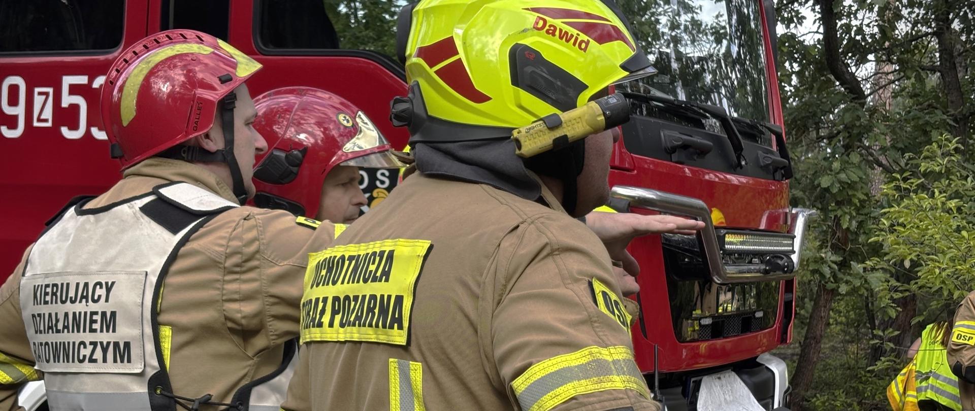 Na zdjęciu widać grupę strażaków stojących przy czerwonym wozie strażackim w lesie. Dwóch z nich nosi czerwone kaski, a jeden z przodu ma żółty kask z napisem "Dawid". Na plecach mają naszywki z napisami "Kierujący Działaniem Ratowniczym" oraz "Ochotnicza Straż Pożarna". Strażacy przygotowują się do działań ratowniczych. W prawym dolnym rogu znajduje się logo z napisem „Fotografia Ratownicza Stargard”.