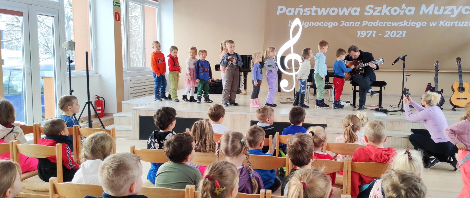 na scenie nauczyciel prezentuje różne rodzaje gitar, przedszkolaki grają na jednej z nich