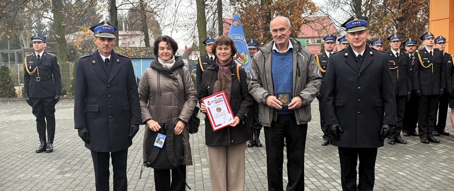 Przed budynkiem Komendy Powiatowej PSP odbywa się uroczysty apel. Stojący przed funkcjonariuszem ludzi otrzymują wyróżnienie. Podniosła atmosfera. W tle widoczne są drzewa w jesiennych barwach oraz fragment nowoczesnego budynku strażnicy.