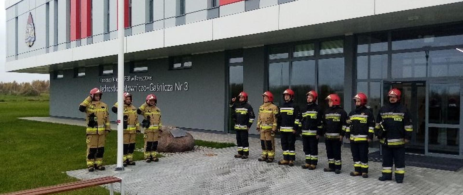 Zdjęcie przedstawia strażaków z Jednostki Ratowniczo-Gaśniczej numer 3 w Rzeszowie w ubraniach specjalnych stojących przed budynkiem strażnicy. Dowódca szyku i poczet flagowy oddają honor, flaga państwowa jest podniesiona na maszcie. 