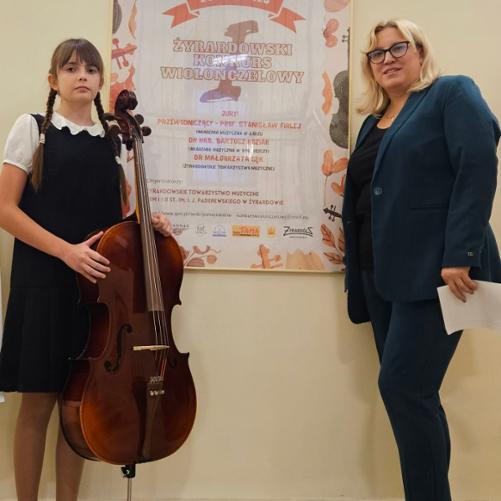 Zdjęcie przestawia Weronikę Krzyżewską i panią Urszulę Właźniak stojące po bokach plakatu reklamującego I Żyrardowski Konkurs Wiolonczelowy. Plakat w kolorach: brązowo, pomarańczowo, czarno, szarym. Na górze plakatu wiolonczela, poniżej informacje dotyczące konkursu.