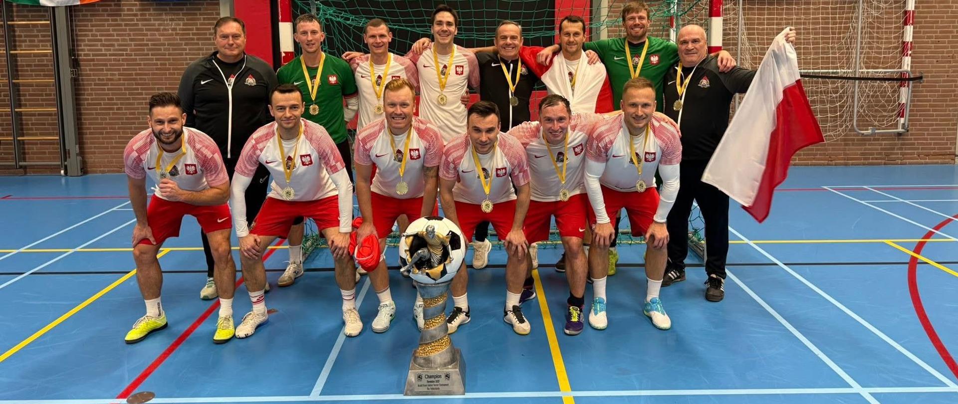 Drużyna Reprezentacji Polski Strażaków pozuje wspólnie przed bramką w hali sportowej. Zawodnicy stoją i kucają w dwóch rzędach, prezentując złote medale i puchar ustawiony przed nimi. W tle widoczne są międzynarodowe flagi zawieszone nad boiskiem.