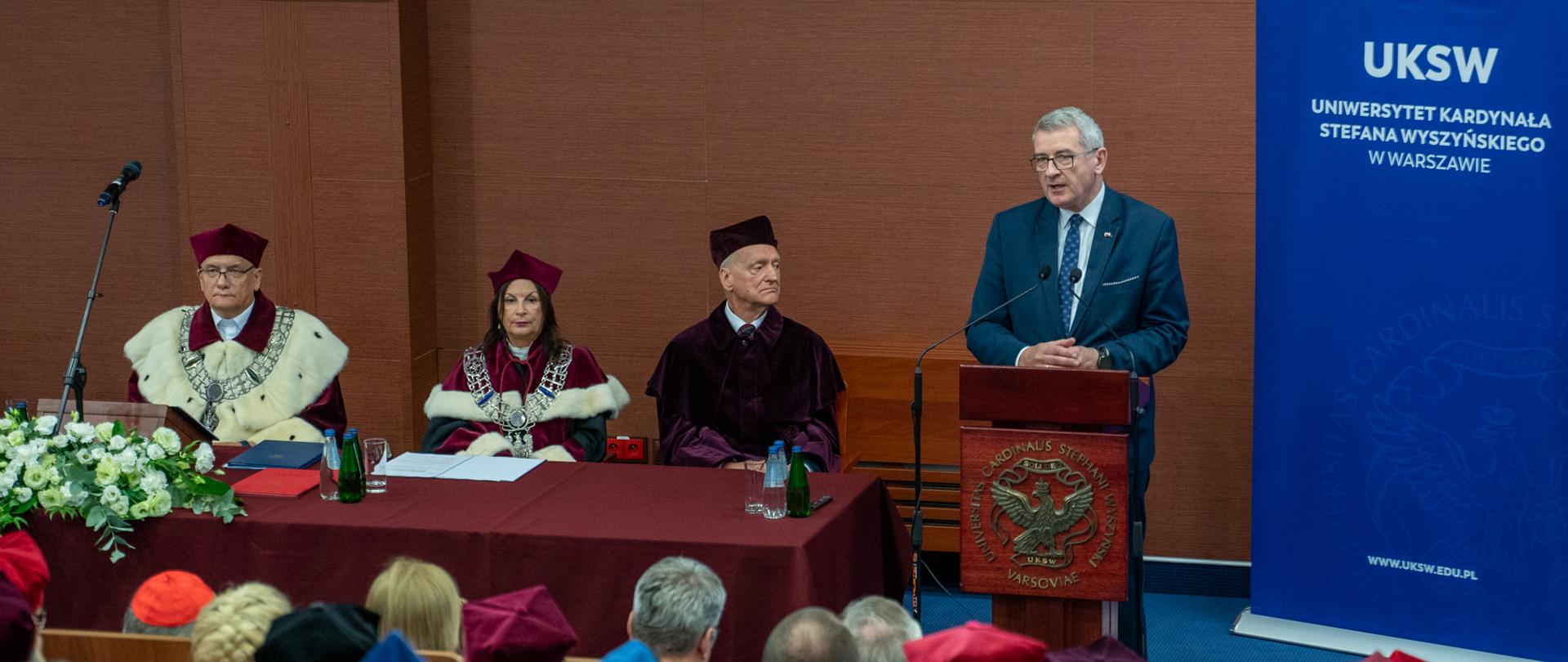 Na sali za mównica stoi wiceminister Murdzek i mówi do mikrofonu, obok niego stół na którym siedzi kilka osób w czerwonych ceremonialnych strojach z gronostajowymi kołnierzami, z boku niebieska ścianka z napisem UKSW.