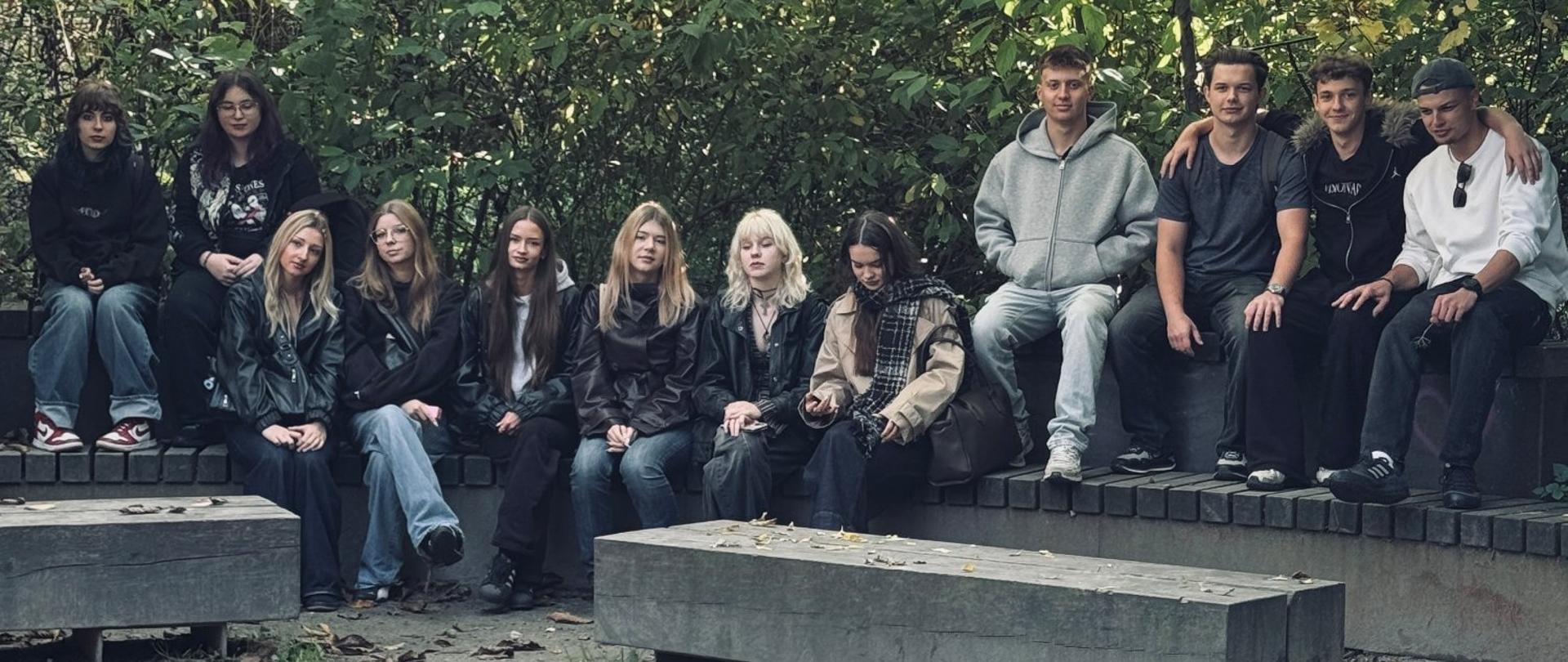 Uczestnicy wycieczki siedzą na półkolistym murku w otoczeniu gęstej zieleni parku. Na pierwszym planie znajdują się betonowe ławki i fragment drewnianego podestu.