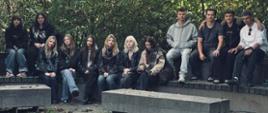 Grupa uczestników wycieczki pozuje do wspólnego zdjęcia na szerokim drewnianym tarasie przy zbiorniku wodnym. Za nimi rozciąga się krajobraz ze skałami i zielenią.