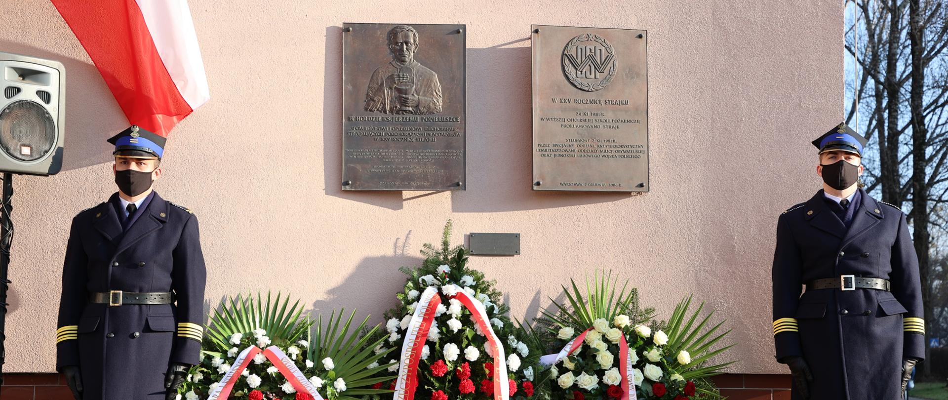 Na ścianie budynku szkoły wiszą dwie tablice upamiętniające ks. J.Popiełuszko oraz uczestników strajku, pod tablicami wiązanki kwiatów a po bokach stoi dwóch strażaków w umundurowaniu wyjściowym