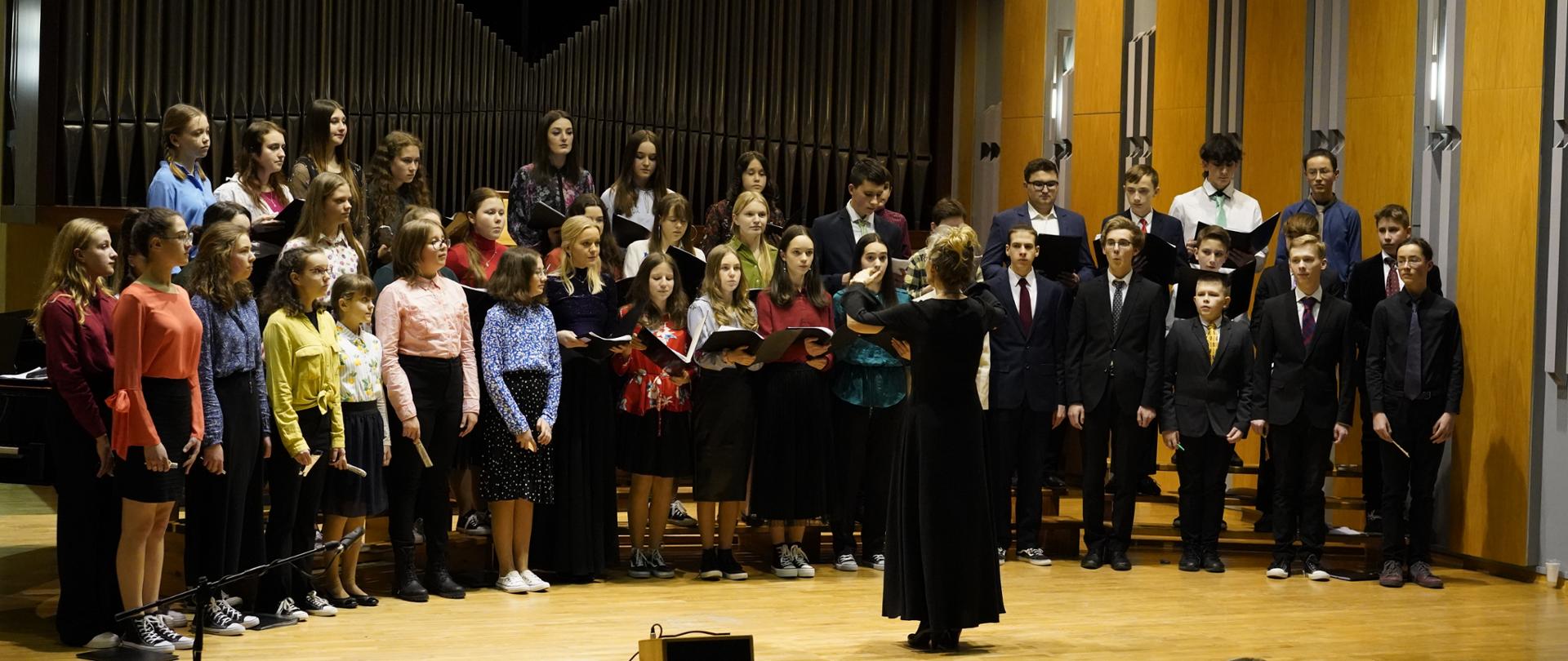 Zdjęcie przedstawia chór szkolny. Chór stoi w trzech rzędach w kolorowych ubraniach. Dzieci w ręku trzymają teczki z nutami. Przed chórem stoi i dyryguje Pani dyrygent. W tle organy, z przodu głośnik i publiczność.