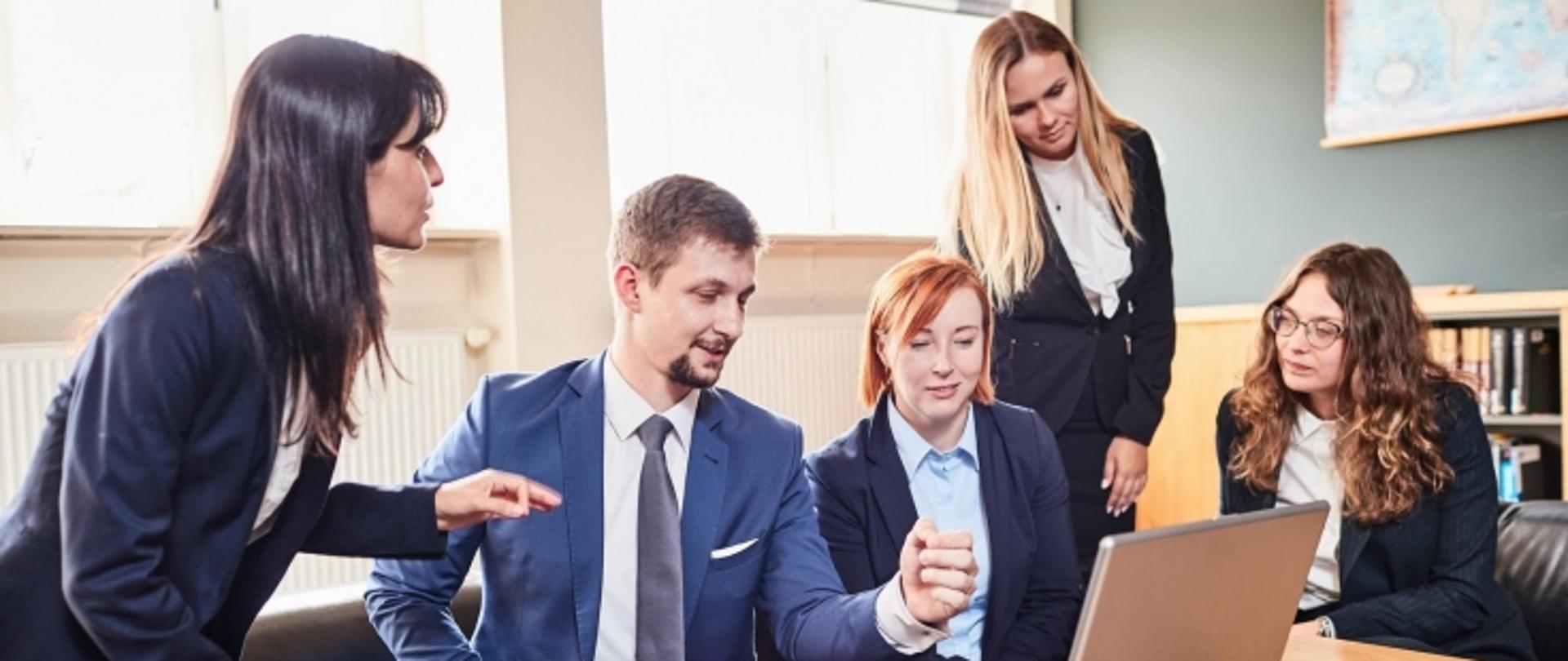 Kilka osób ubranych w sposób formalny siedzi na kanapie i ogląda coś co jest wyświetlane na laptopie