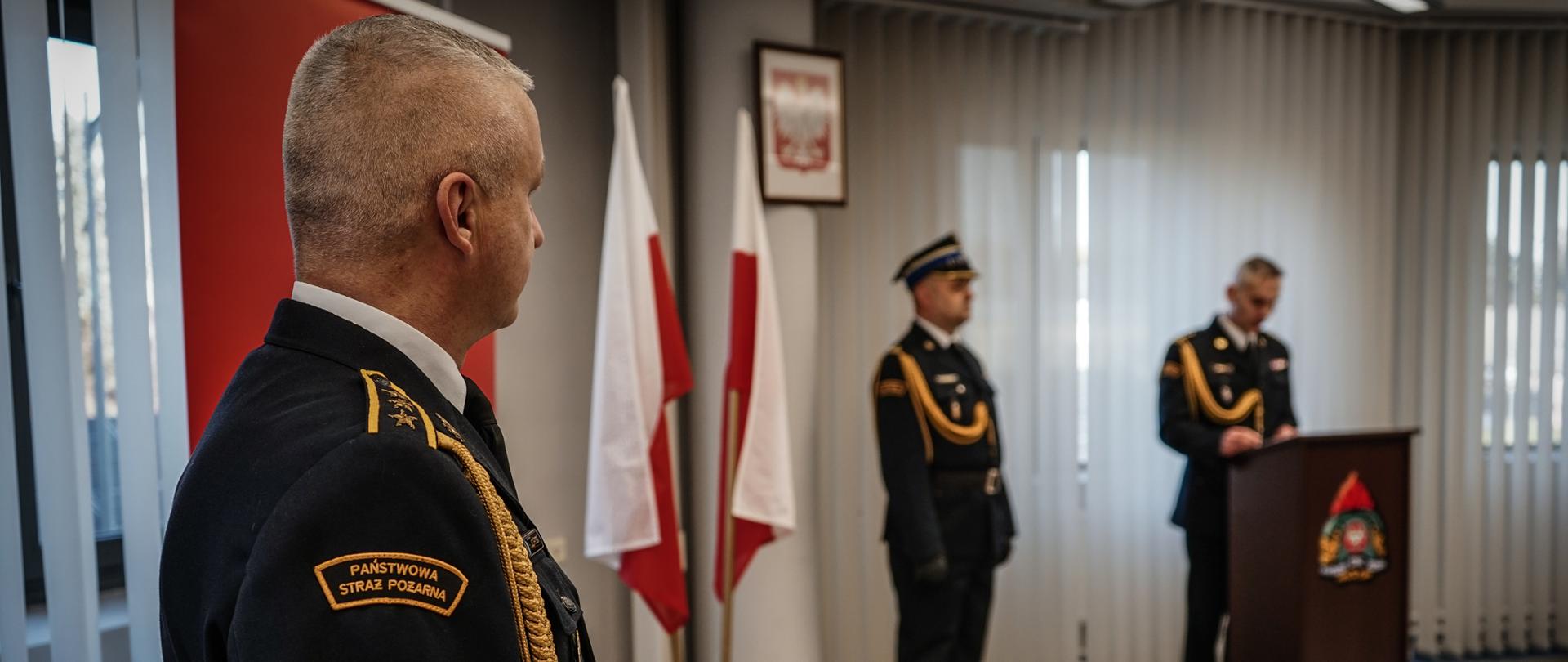 Zdjęcie zrobione w pomieszczeniu strażnicy na tle godła Rzeczpospolitej Polskiej, oraz dwóch Flag Rzeczpospolitej Polskiej. Na pierwszym planie strażak oraz w oddali dwóch strażaków w umundurowaniu galowym, jeden z nich stoi przy mównicy.