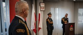 Zdjęcie zrobione w pomieszczeniu strażnicy na tle godła Rzeczpospolitej Polskiej, oraz dwóch Flag Rzeczpospolitej Polskiej. Na pierwszym planie strażak oraz w oddali dwóch strażaków w umundurowaniu galowym, jeden z nich stoi przy mównicy.