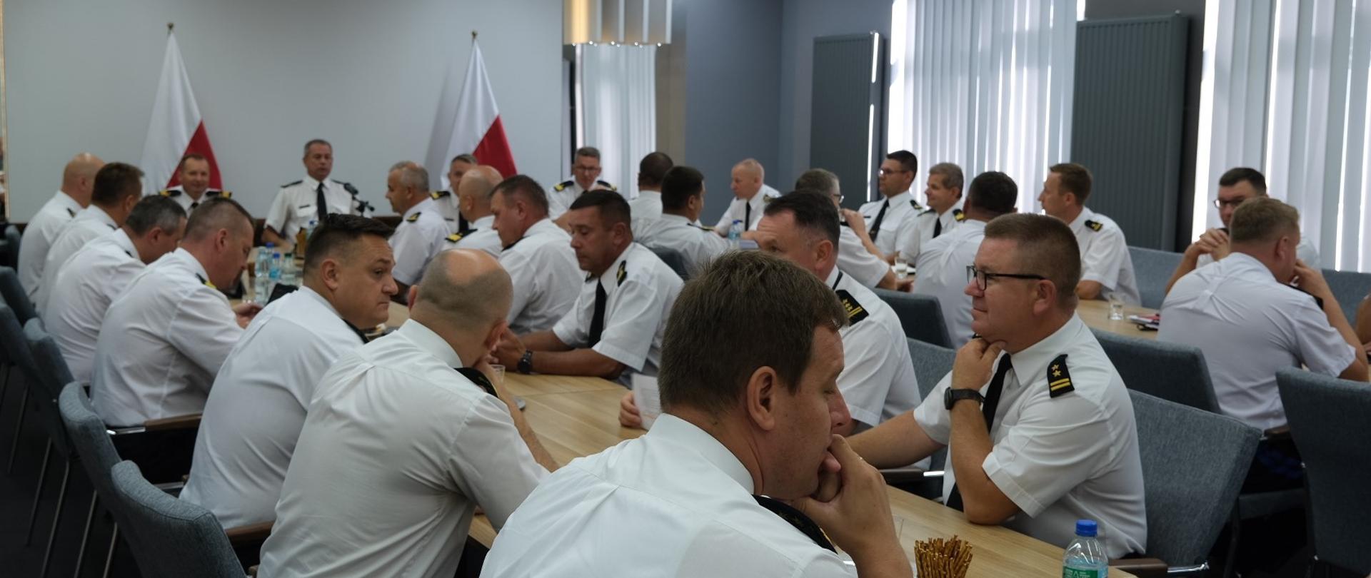 strażacy w białych koszulach z dystynkcjami i w czarnych krawatach siedzą przy stołachg, odprawa, w tle swie biało czerwone flagi