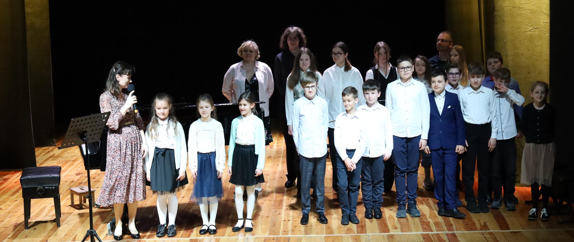 Fotografia wspólna wszystkich wykonawców koncertów dla uczniów szkół podstawowych. Ustawieni w rzędzie na scenie MOK w Głubczycach, przed oraz z prawej strony obok fortepianu.