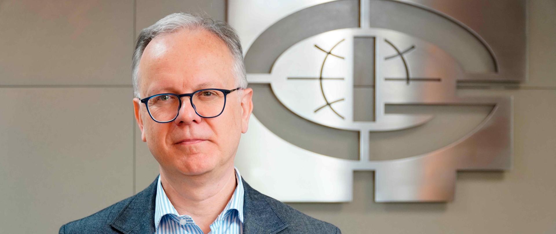 Prof. Paweł Rowiński stoi na tle szarej ściany z logiem IGF.