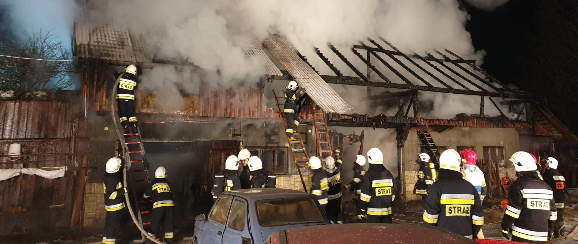 na zdjęciu są zabudowania gospodarcze które się palą a strażacy stojąc na drabinach gaszą je część budynku ma spalony całkowicie dach
