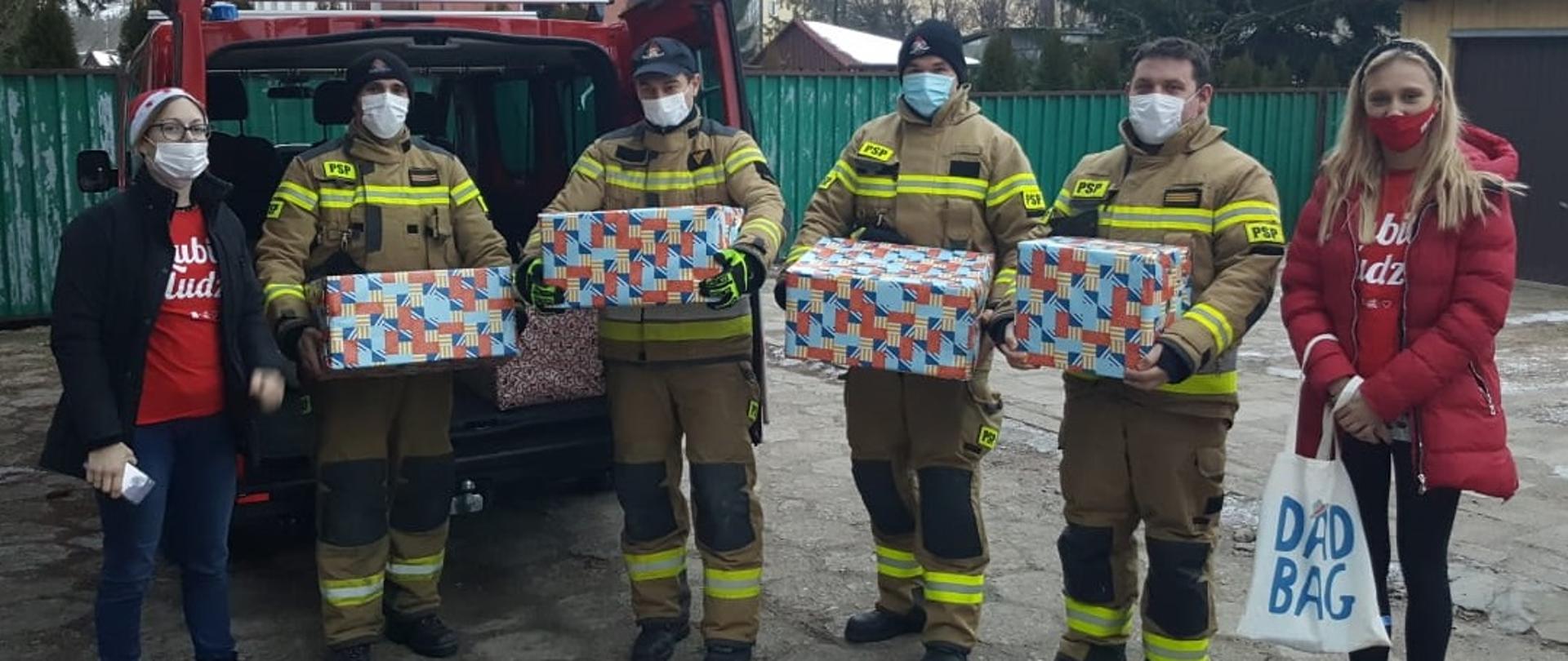 Czterech funkcjonariuszy i dwie wolaontariuszju stoją na podórku przy samochodzie strażackim trzymając paczki do przekazania, Za nimi widac blok. 