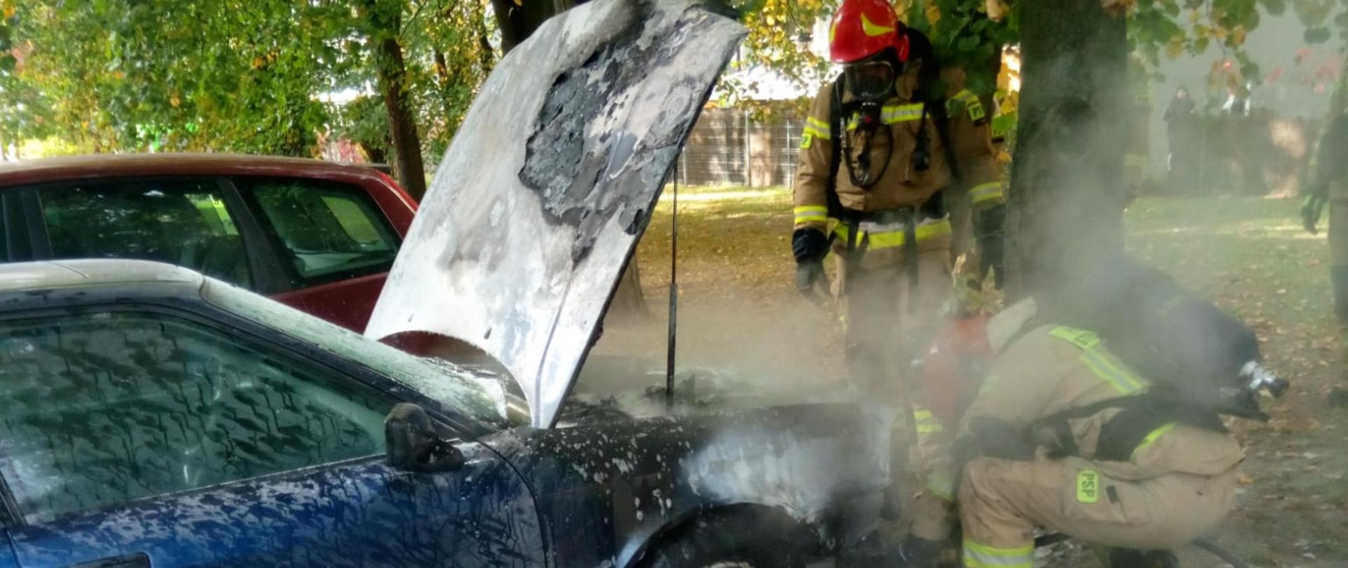 Zdjęcie przedstawia samochód osobowy, w którym zapaliła się komora silnika. Dwóch ratowników w aparatach ochrony dróg oddechowych prowadzi działania podając do przodu piane ciężką. Maska auta jest otwarta. 