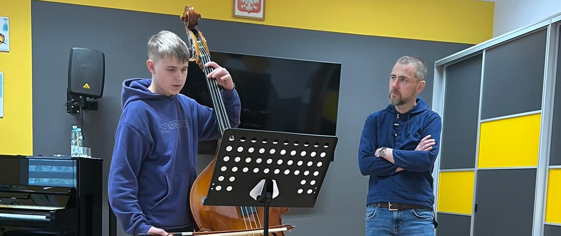 Chłopiec w niebieskiej bluzie gra na kontrabasie, stojąc w sali muzycznej z drewnianą podłogą. Obok niego stoi nauczyciel, z założonymi rękami, obserwujący chłopca. W tle widać pianino, głośnik, telewizor i tablicę z herbem Polski nad ekranem. Ściany są w odcieniach szarości i żółci.