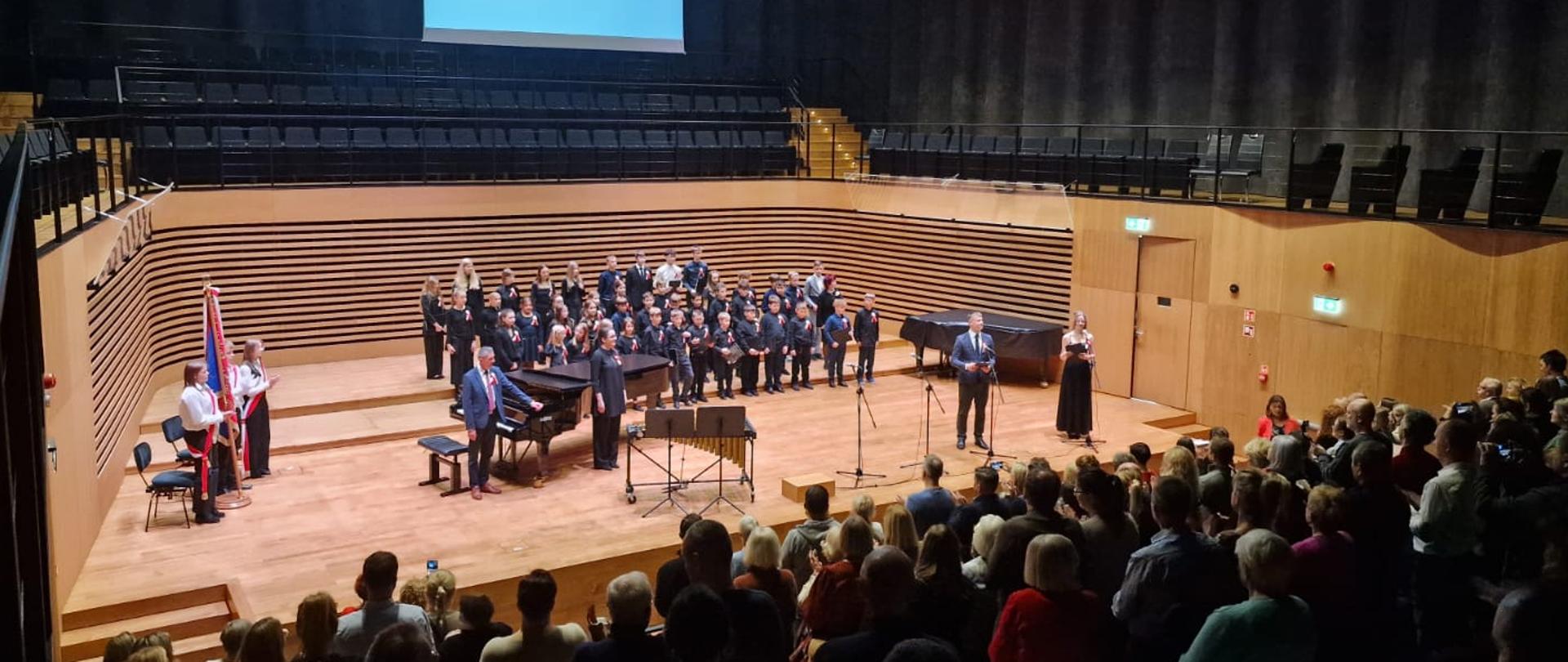 Na estradzie stoi dziewczyna i chłopak, kobieta i mężczyzna oraz chóry i poczet sztandarowy, z przodu widoczna stojąca publiczność.