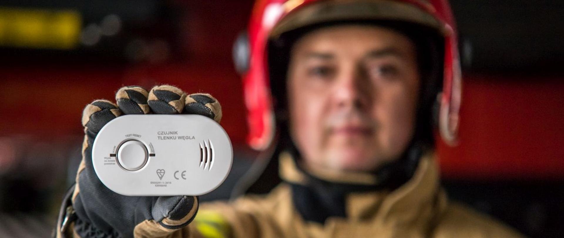 Strażak w ubraniu specjalnym koloru piaskowego w czerwonym hełmie trzyma w wysuniętej ręce czujnik tlenku węgla. Funkcjonariusz stoi na tle samochodu pożarniczego.