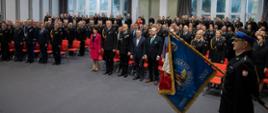 Na sali kilkaset osób - funkcjonariuszy w strojach galowych i gości uroczystości. Po prawej stronie poczet sztandarowy pochyla sztandar.