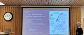 Kobieta prowadzi prezentację w sali konferencyjnej. Na ekranie wyświetlony jest slajd dotyczący funduszy EOG i Funduszy Norweskich oraz zwiększania odporności na katastrofy.