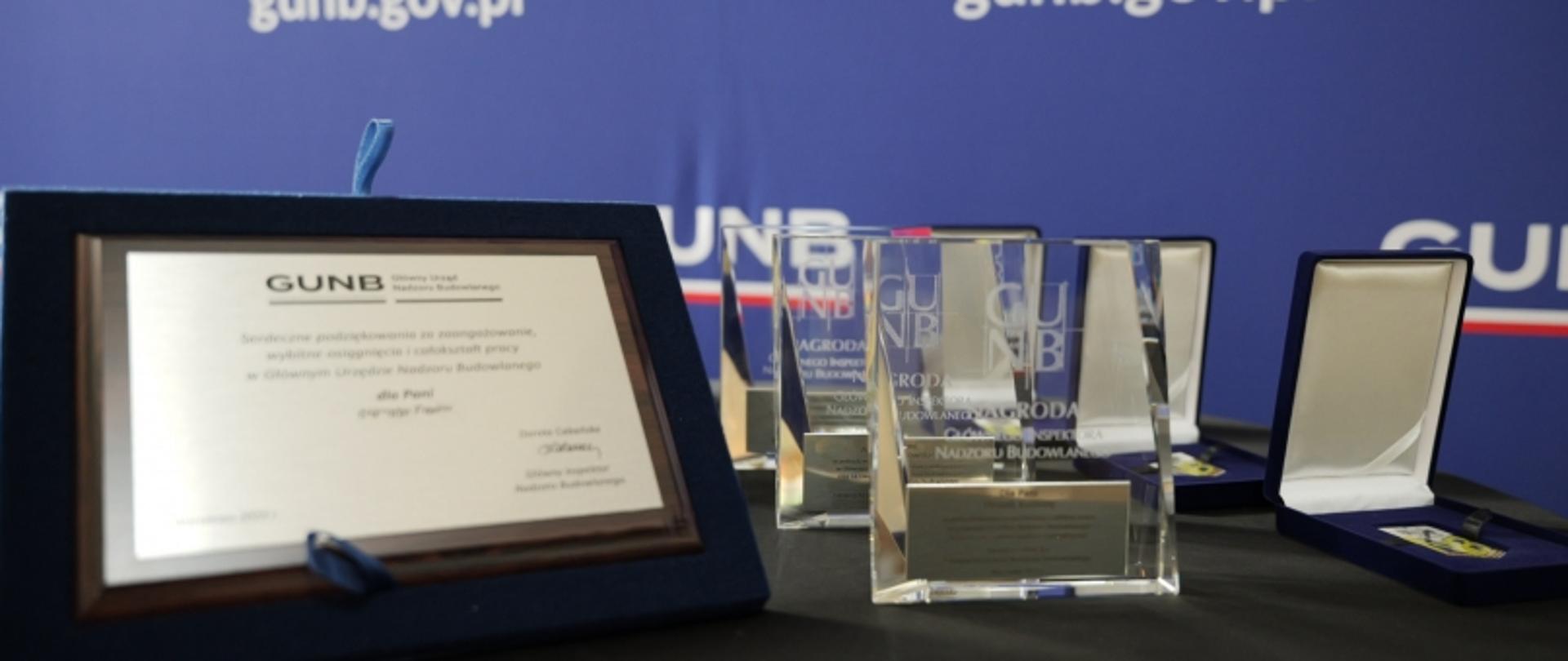 Zestaw szklanych statuetek i oprawionych dyplomów z podziękowaniami od GUNB, ustawionych na stole przed tłem z logo urzędu.