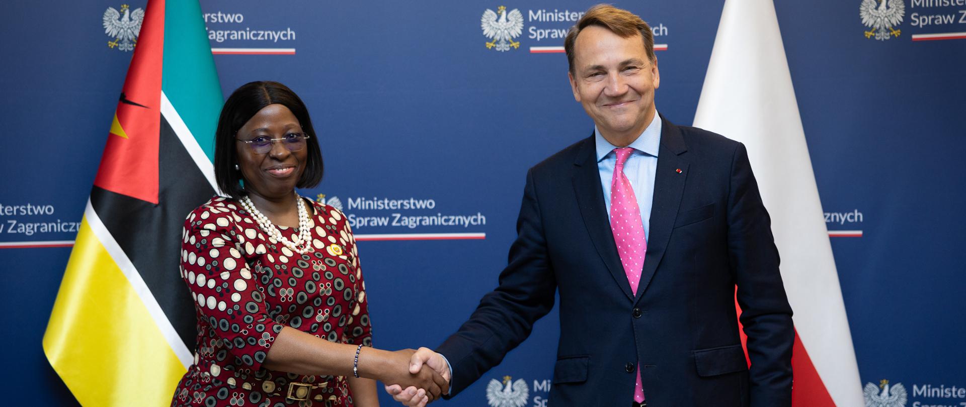 Maria Manuela dos Santos Lucas and Radosław Sikorski shake hands against a navy blue backdrop featuring the logos of the Polish Ministry of Foreign Affairs and the flags of Mozambique and Poland. Both are smiling for the camera. The photograph documents the official meeting.