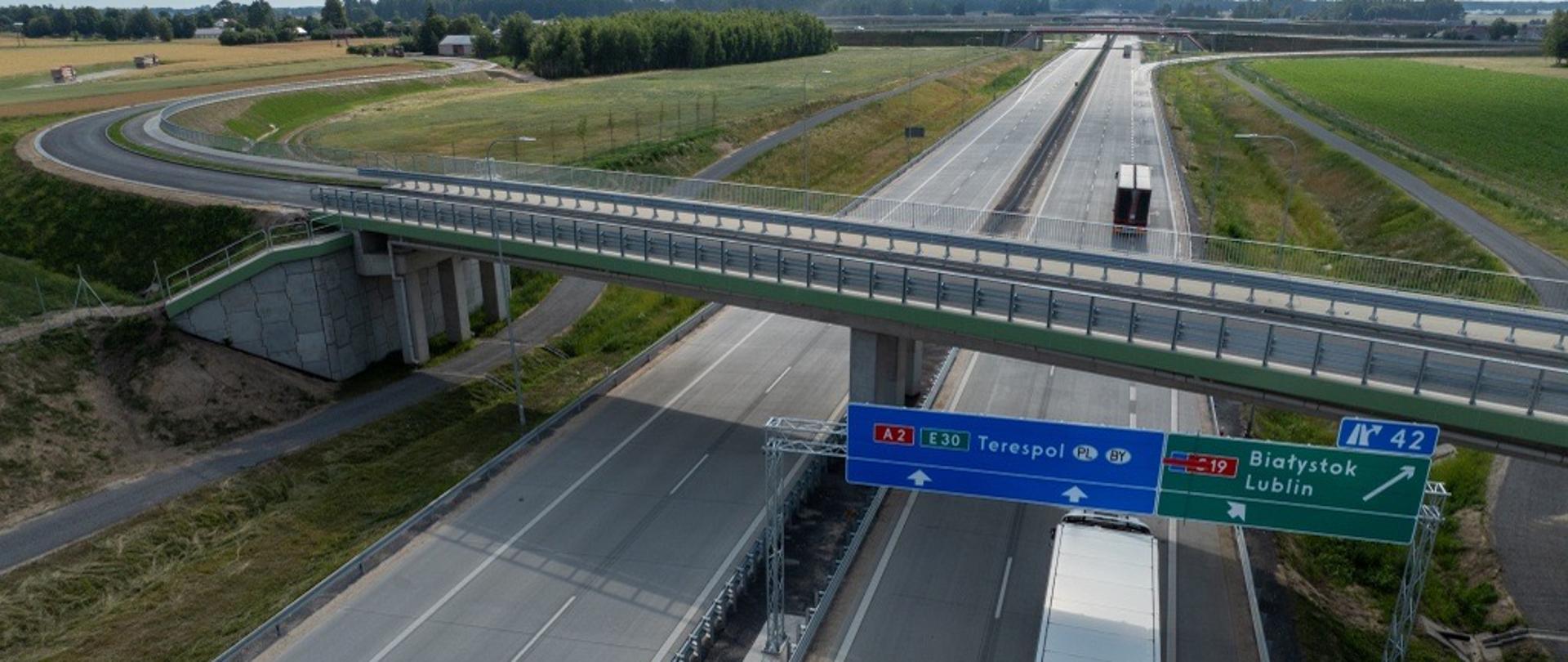 Dwujezdniowa autostrada A2 widziana z góry, z wiaduktem przebiegającym nad jezdniami i ciężarówką jadącą prawym pasem. Nad drogą znajdują się znaki kierunkowe: A2/E30 na Terespol oraz zjazd w kierunku drogi S19 na Białystok i Lublin. Wokół rozciągają się pola i tereny zielone.
