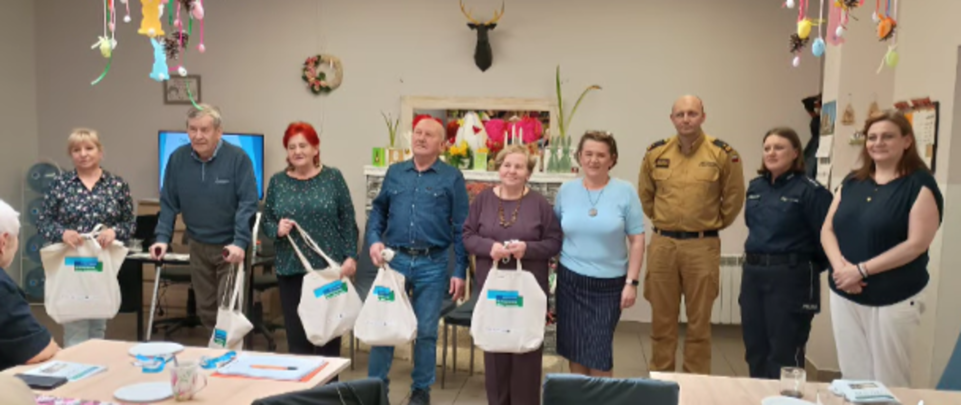 Na zdjęciu widać grupę kilku osób stojących w pomieszczeniu Klubu Senior+. Wśród nich są seniorzy oraz przedstawiciele służb mundurowych – policji i straży pożarnej. Część uczestników trzyma w rękach torby z materiałami lub upominkami. Wszyscy ustawieni są w jednym rzędzie, pozując do wspólnego zdjęcia.
Sala jest udekorowana kolorowymi ozdobami wiszącymi pod sufitem, co nadaje przestrzeni przyjazny i świąteczny charakter. W tle znajdują się stoły, krzesła oraz elementy wyposażenia świetlicy. Atmosfera spotkania wydaje się spokojna i życzliwa, a uczestnicy wyglądają na zadowolonych z udziału w wydarzeniu.