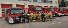 funkcjonariusze PSP stoją zgrupowani przed samochodami pożarniczymi, w tle budynek Komendy PSP