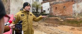 Strażak, który opowiada o realizacji inwestycji - stoi na placu rozbiórki, w tle mur średniowieczny Torunia. Z przodu widać dziennikarzy.