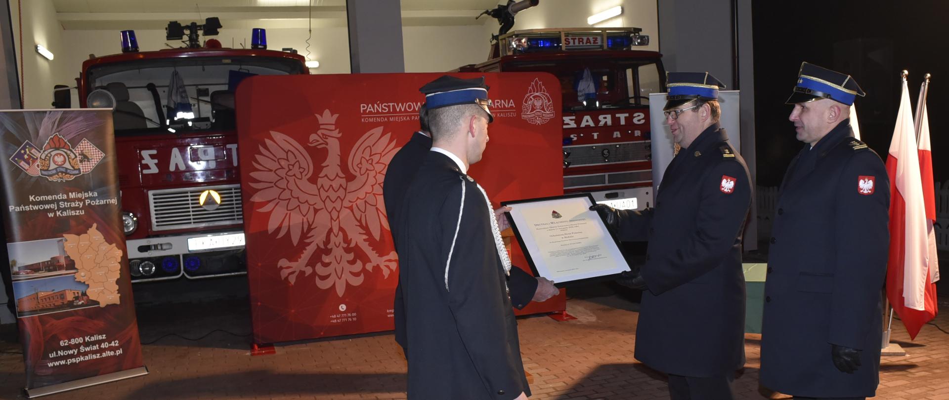 dwóch strażaków wręcza innym strażakom ramkę z dokumentem , w tle samochody strażackie i duża tekstylna ścianka z orłem
