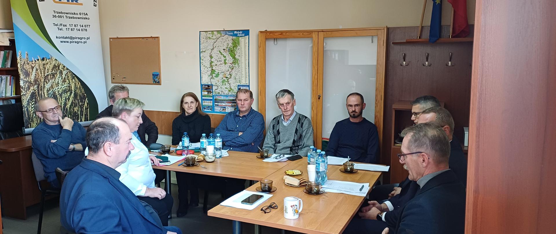 Grupa ludzi siedząca przy stole na którym rozmieszczone są butelki z wodą. Pomieszczenie jest jasne. Po lewej stronie stoi baner z logiem instytucji. Po prawej stronie są brązowe szafy.