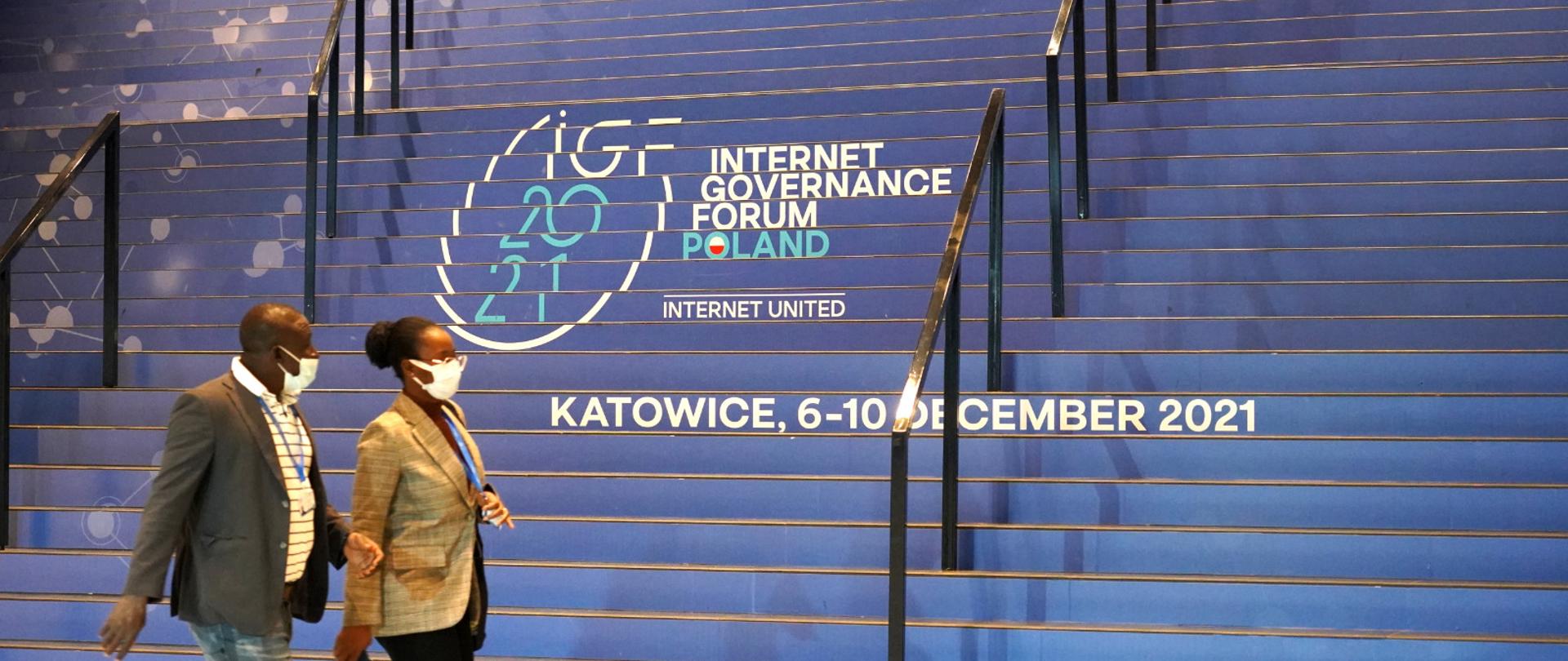 Dwoje uczestników Szczytu przechodzi obok schodów, na których widoczne jest logo IGF 2021 i napis Katowice, 6-10 grudnia 2021.