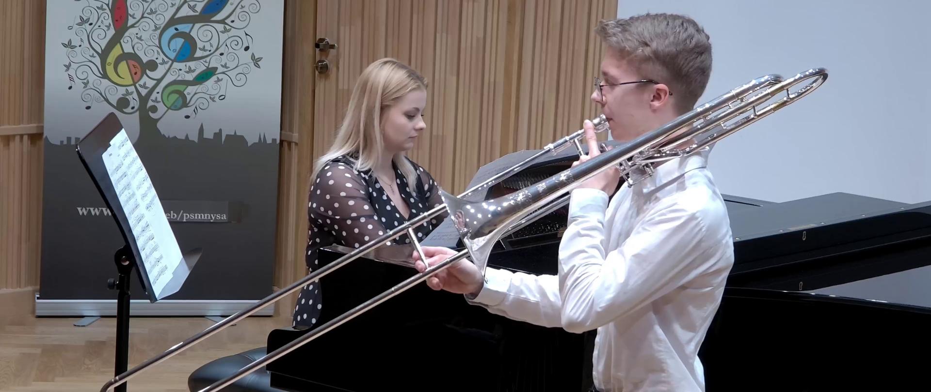Zdjęcie z koncertu uczniów z klas instrumentów dętych PSM I i II stopnia w Nysie - Sala Koncertowa. Na pierwszym planie puzonista z instrumentem, oraz pianistka grająca na czarnym fortepianie. w tle baner PSM Nysa.