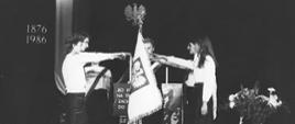 Na zdjęciu widzimy uroczystość, która odbyła się 24 października 1986 r. z okazji 110-lecia szkoły na scenie Teatru im. Stanisława Ignacego Witkiewicza w Zakopanem. Na pierwszym planie widzimy ucznia Józefa Brzuchacza trzymającego przed sobą sztandar szkoły. Po lewej i prawej stronie stoją uczniowie szkoły - dwie uczennice i dwóch uczniów, którzy trzymając uniesione swoje prawe ręce składają uczniowskie ślubowanie. Uczniowie ubrani są odświętnie w białe koszule i czarne spodnie, dziewczyny czarne spódnice. Po lewej stronie na ścianie wisi w pionie narodowa flaga. Widnieje również data: 1876-1986. Z tyłu widzimy również godło państwowe - orła bez korony wyrzeźbionego z drewna. Po prawej stronie widzimy ceramiczny wazon w którym włożone są goździki. Zdjęcie jest czarno-białe ale wiemy że kwiaty w wazonie są w barwach narodowych, biało-czerwone. Autorem fotografii jest: Stanisław Momot 