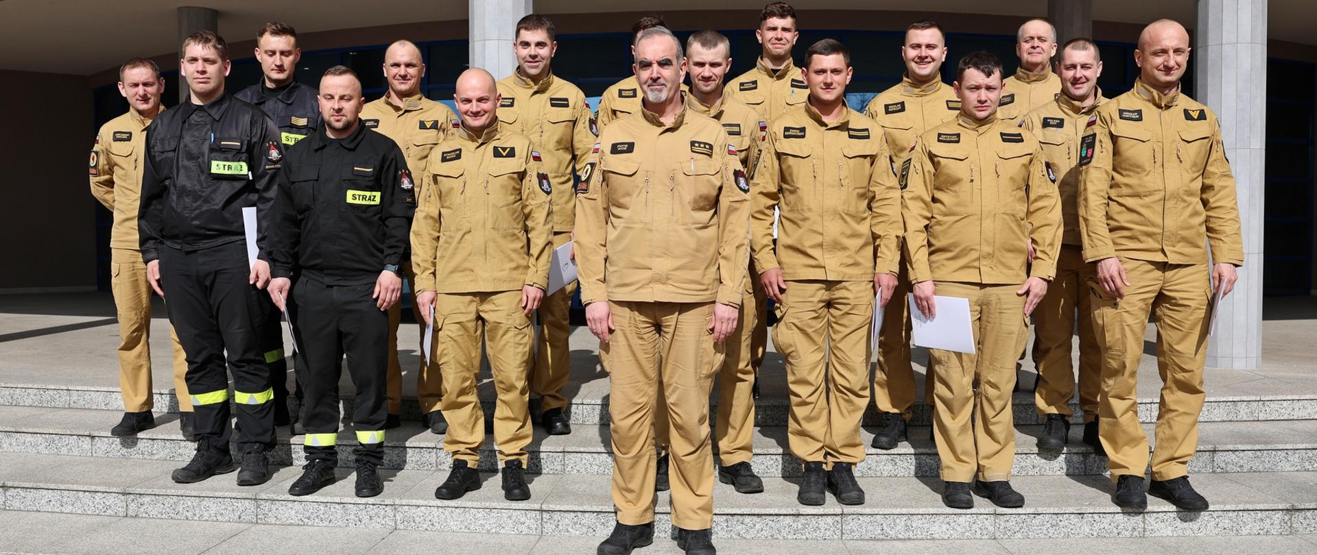 Pamiątkowe zdjęcie grupowe funkcjonariuszy PSP będących uczestnikami szkolenia dla kierowców pojazdów uprzywilejowanych (14 osób) w towarzystwie Zastępcy Komendanta Centralnej Szkoły PSP oraz dwóch funkcjonariuszy CS PSP. Grupa ustawiona jest na schodach przed budynkiem