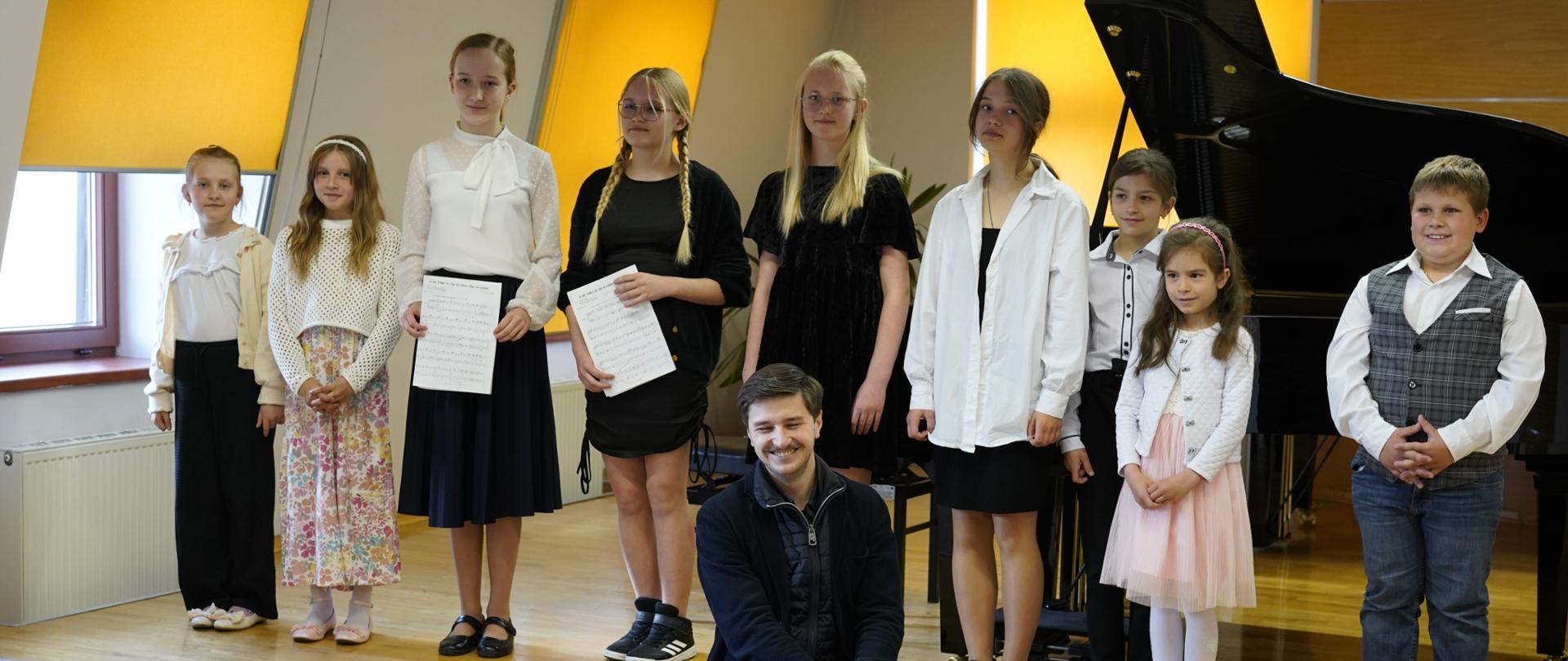 Na zdjęciu znajduje się grupa młodych uczestników audycji duetów fortepianowych stojących na scenie w sali kameralnej. Jest ich dziesięcioro – osiem dziewczynek i dwóch chłopców – ubranych elegancko, jak na występ muzyczny przystało. Część dzieci trzyma w rękach nuty. W tle widać fortepian oraz pomarańczowe rolety w oknach. Na pierwszym planie, przed sceną, siedzi uśmiechnięty młody mężczyzna – nauczyciel prowadzący audycję. Trzyma w ręku bordową teczkę i pozuje do wspólnego zdjęcia z dziećmi. Atmosfera jest radosna i uroczysta, a dzieci wyglądają na dumne i zadowolone ze swojego występu.