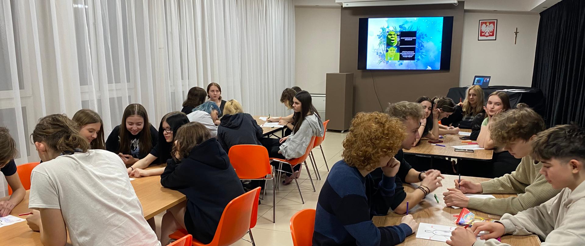 Przy każdym z czterech stołów siedzi po kilkoro dziewcząt i chłopców. Na stolikach leżą kartki i pisaki, niektórzy trzymają je w dłoniach. Siedzą na pomarańczowych krzesłach. Na ścianie na wprost wisi duży telewizor, a na ekranie na niebieskim tle widać znajdujące się jedna nad drugą zielone postaci bajkowego Shreka. Obok na czarnym tle białe napisy - niewyraźne. Po prawej stronie od telewizora na ścianie wisi krzyż i godło Polski.