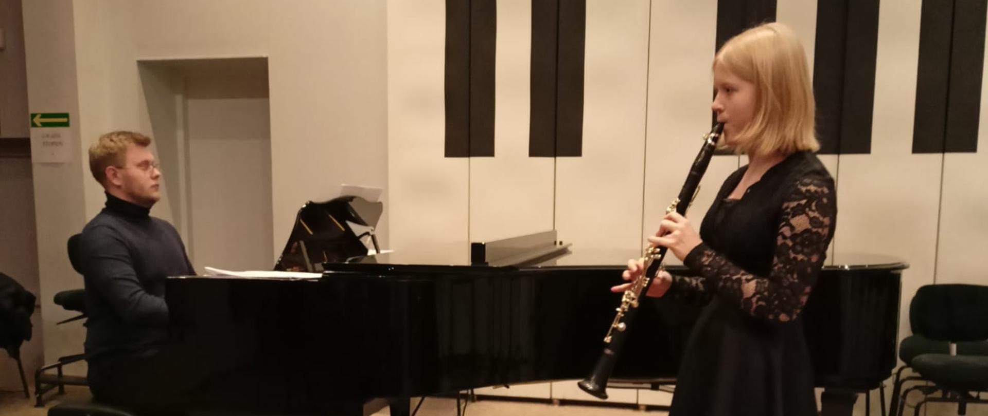 Nastolatka stojąc na estradzie auli PSM gra na klarnecie, za nią po lewej stronie na fortepianie gra mężczyzna.