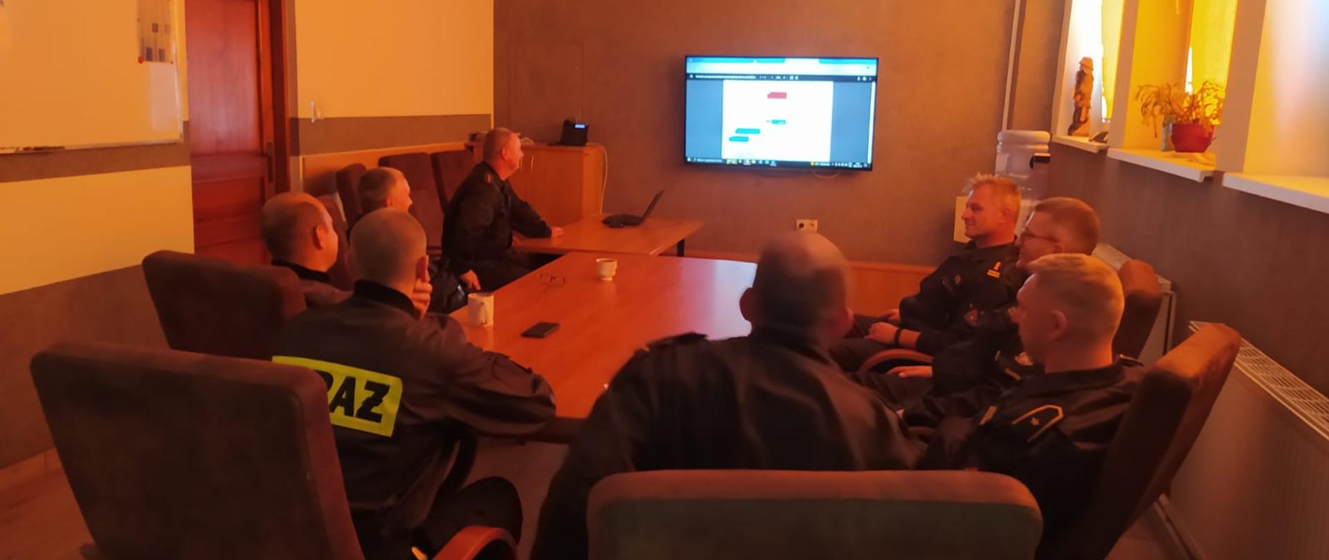 Pomieszczenie w tle telewizor stół przy stole siedzą strażacy (osoby)
