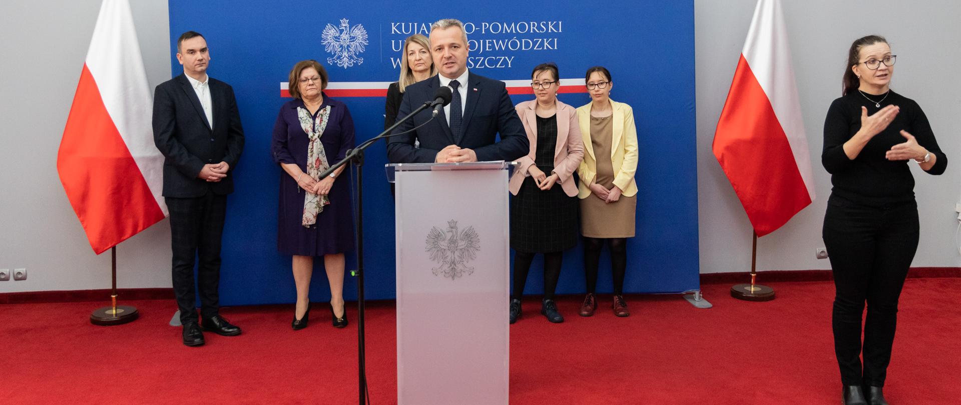 Jeden mężczyzna stoi przed mównicą i wypowiada się. Za nim stoją cztery kobiety i mężczyzna, obok jedna kobieta