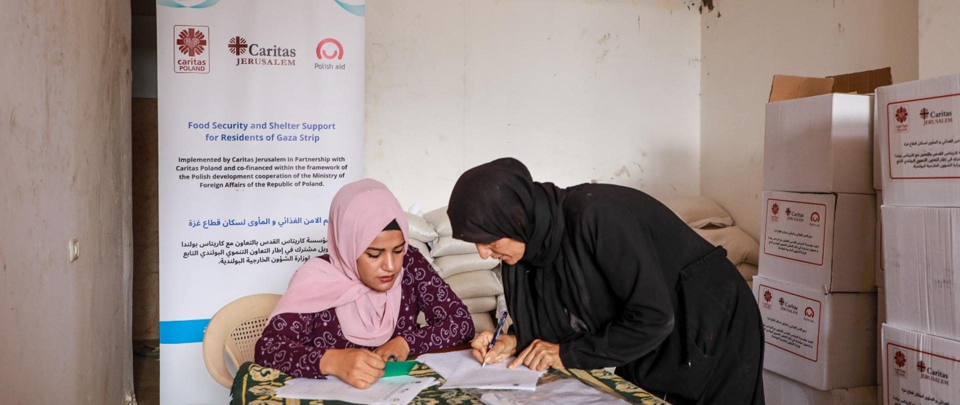 Jedna kobieta z głową zakrytą różową chustą siedzi przy stole, druga kobieta również z głową zakrytą czarną chustą pochyla się nad stołem i pisze na kartce papieru. W tle jest plakat z logo Polskiej pomocy i Caritas oraz białe pudełka. 